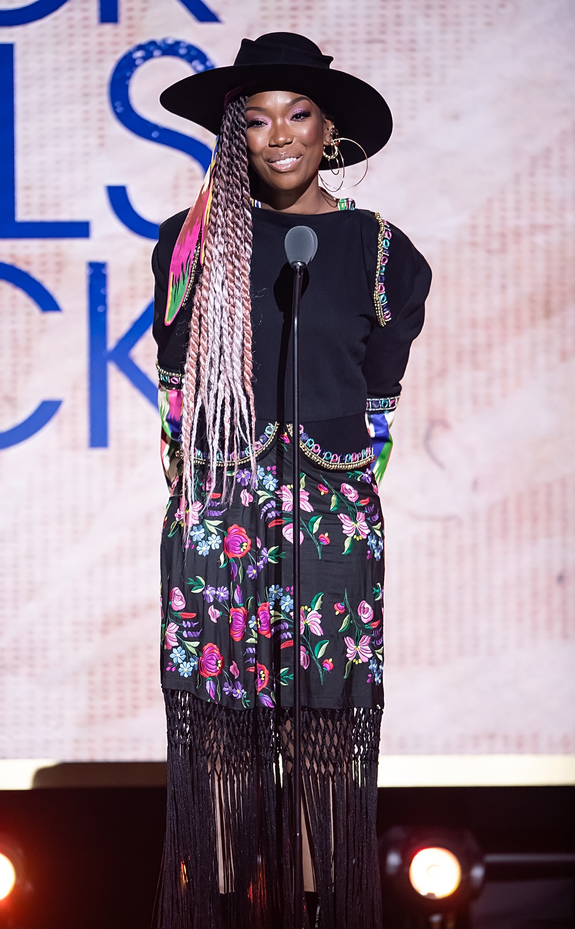 Brandy wearing a floral print skirt, long-sleeved top, and wide-brimmed hat as she speaks onstage