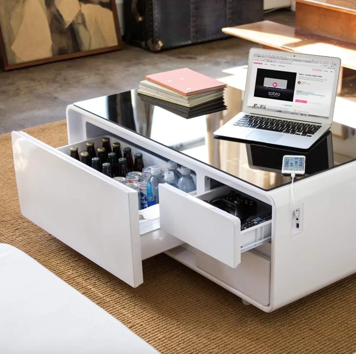 a white coffee table with refrigerated drawers filled with drinks and a phone and a laptop on top plugged in to the built-in outlet