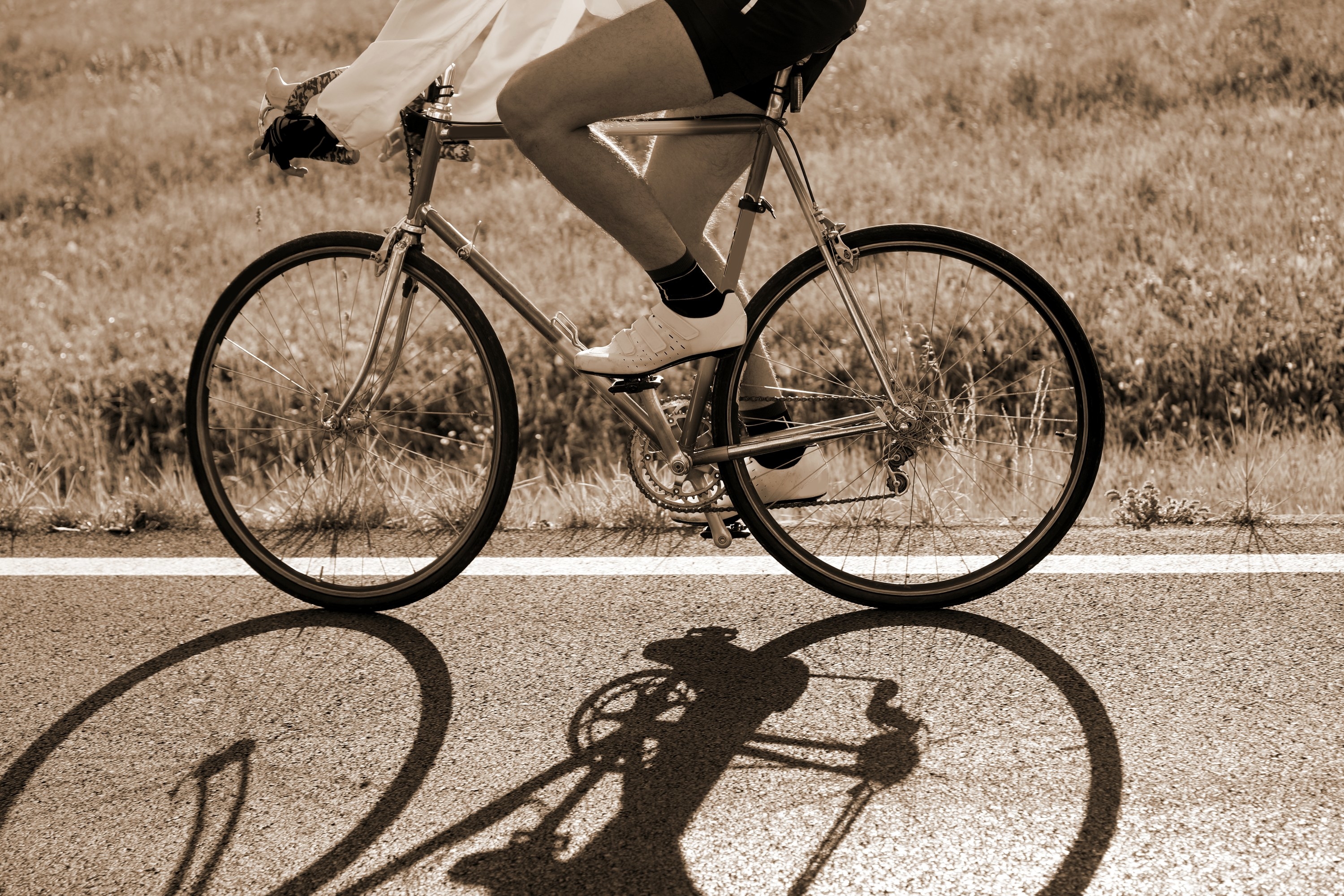 person biking on the side of a road, with grass in the background and their shadow in the foreground