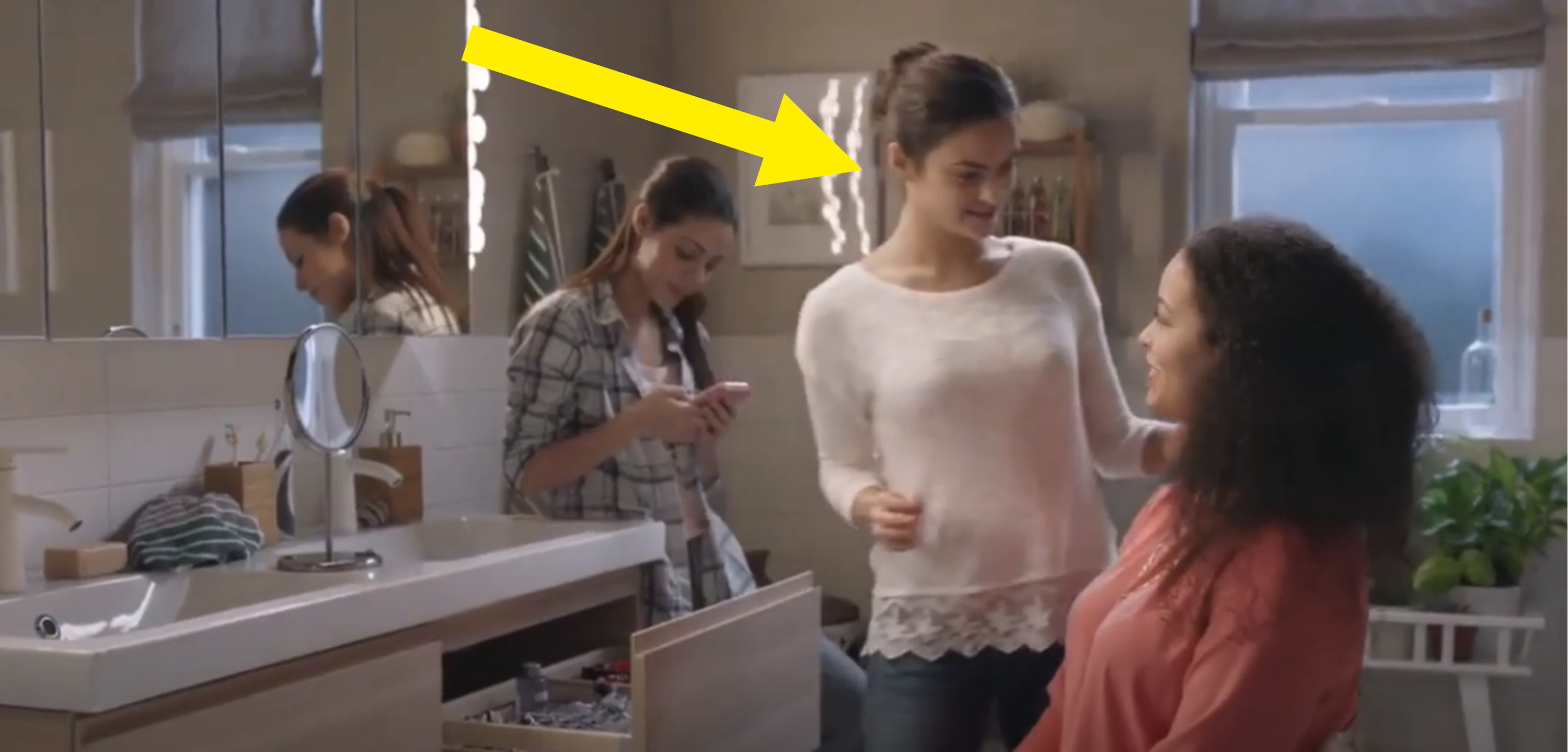 an arrow pointing to Camila in a bathroom with 2 other women for the commercial