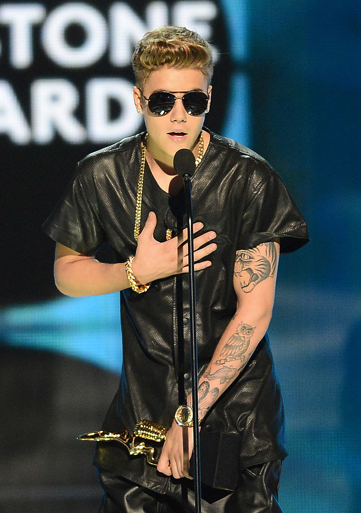 Justin in sunglasses standing in front of a microphone and holding an award in one hand and putting the other hand on his chest