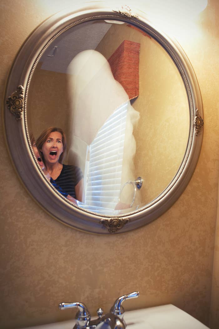 A frightened woman looking at a ghostly figure in her bathroom mirror