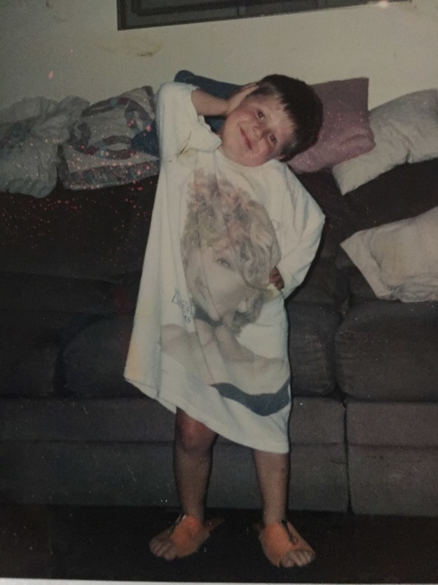 A toddler standing in front of a couch and posing in a long Tina Turner T-shirt