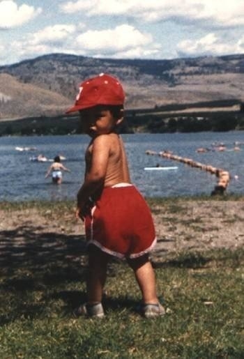 A toddler standing on the grass and wearing a baseball cap and baggy shorts and looking behind them at the camera