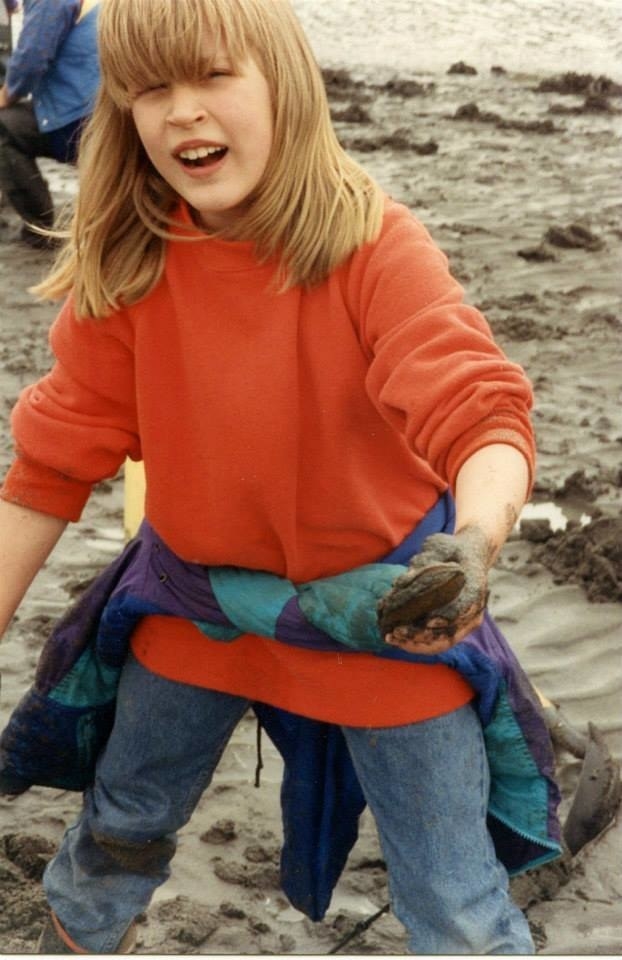 A young girl with bangs standing on sand and wearing jeans and a sweatshirt