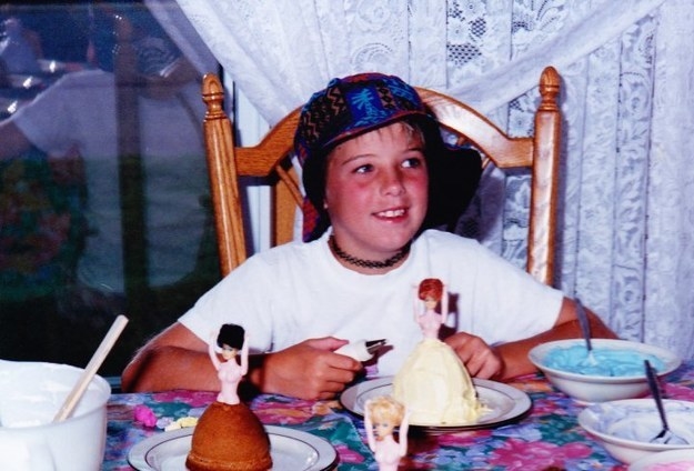 Young person wearing a choker, hat, and T-shirt and sitting at a table with Barbie figures on plates on it