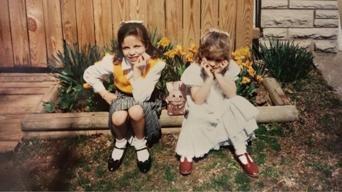 Two girls sitting by plants and both wearing dresses; one is smiling and the one on the right is resting her face in her hands