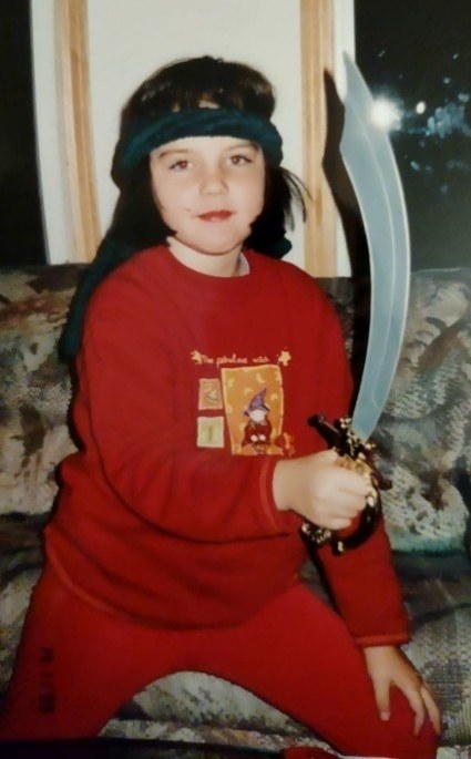 Young girl in sweatpants and sweatshirt, lunging and holding a sword in front of a couch
