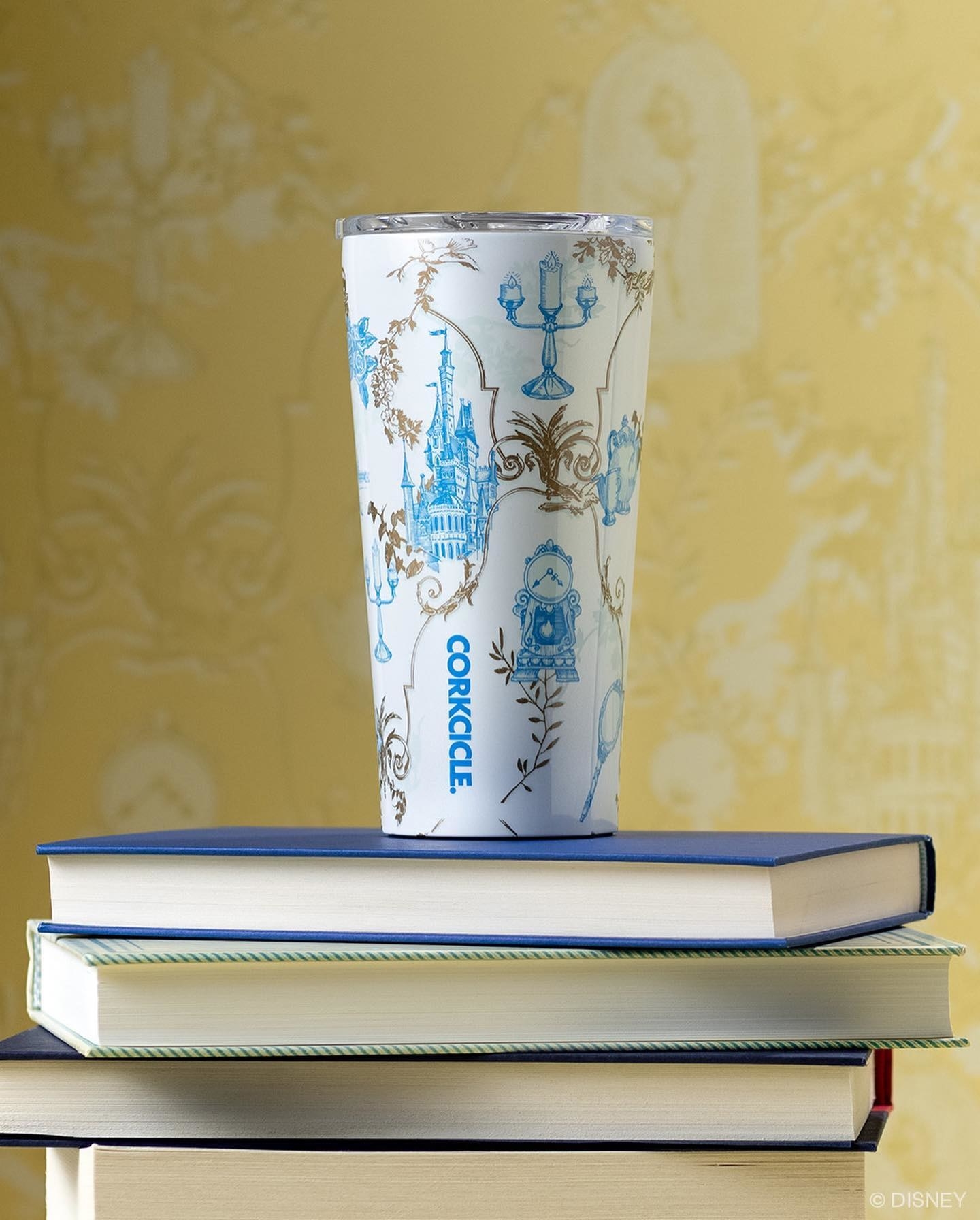 a white corkcicle cup with a light blue and gold beauty and the beast-inspired pattern on it sitting on top of books
