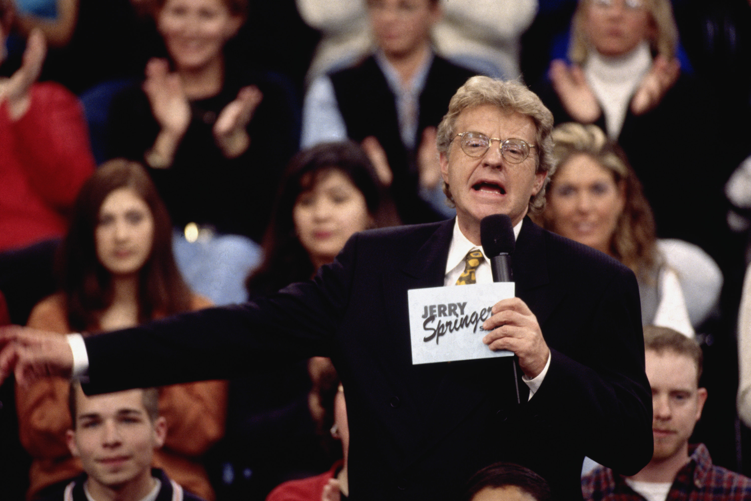 Jerry Springer pointing to something off screen as he talks into the microphone with the audience behind him on &quot;the Jerry Springer Show&quot;
