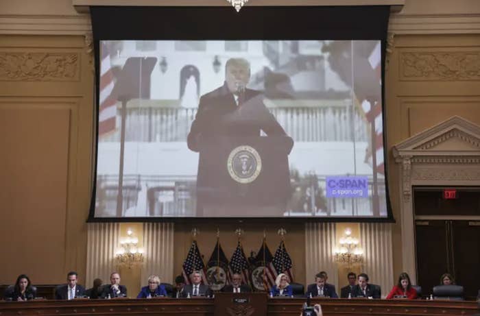 Above a panel of Congress members in suit, a huge projector screen shows Trump speaking at a podium on jan 6