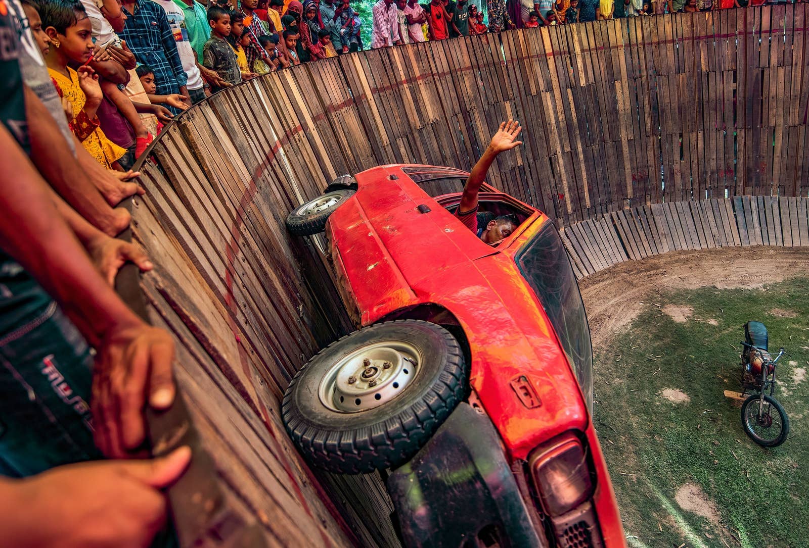 A man waves at the crowd from a red car that is driving around the &quot;Well Of Death,&quot; vertically to the ground.