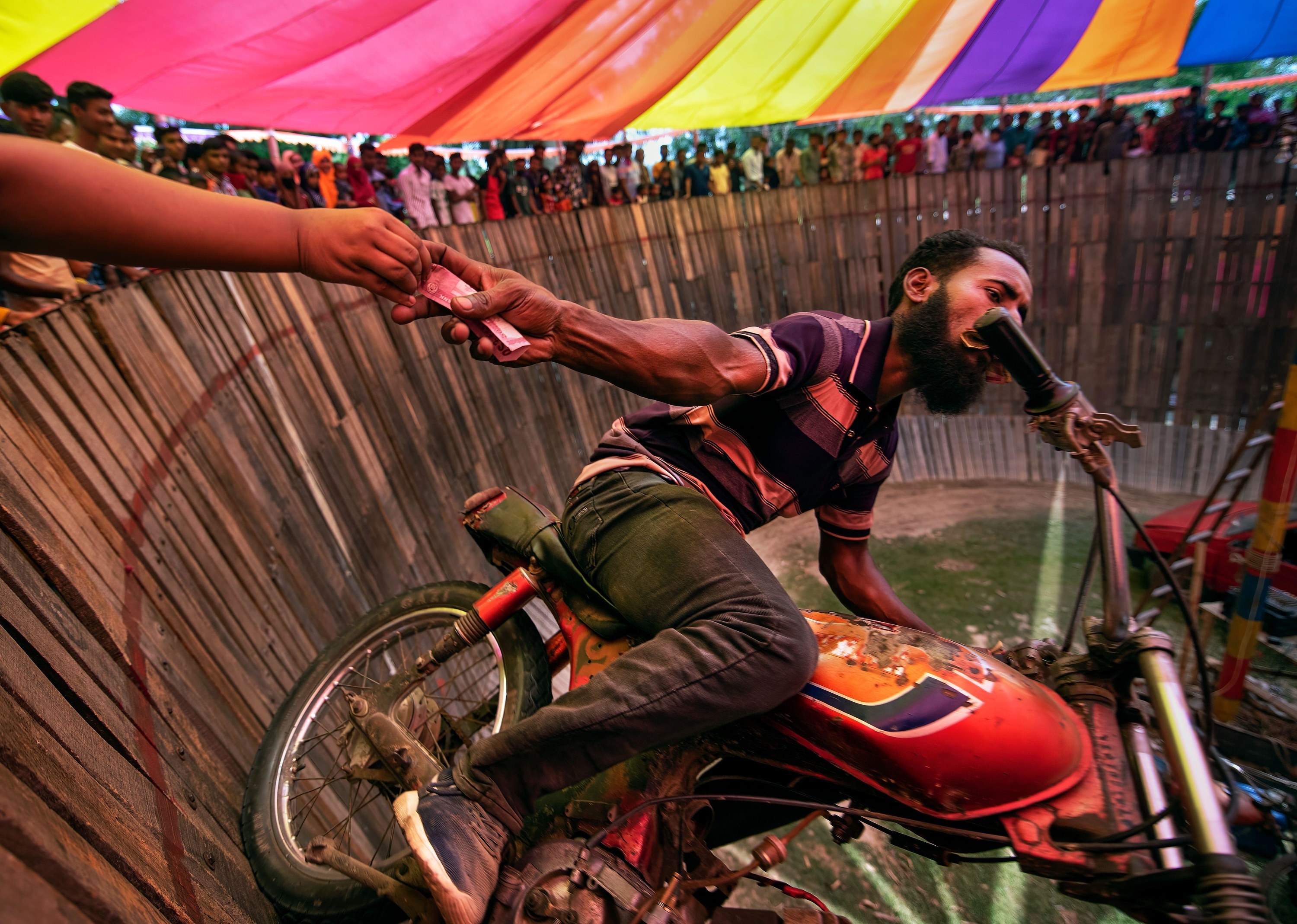 A man riding a motorcycle on the &quot;Well Of Death&quot; grabs a tip offered by an audience member as he rides past, vertically to the ground.