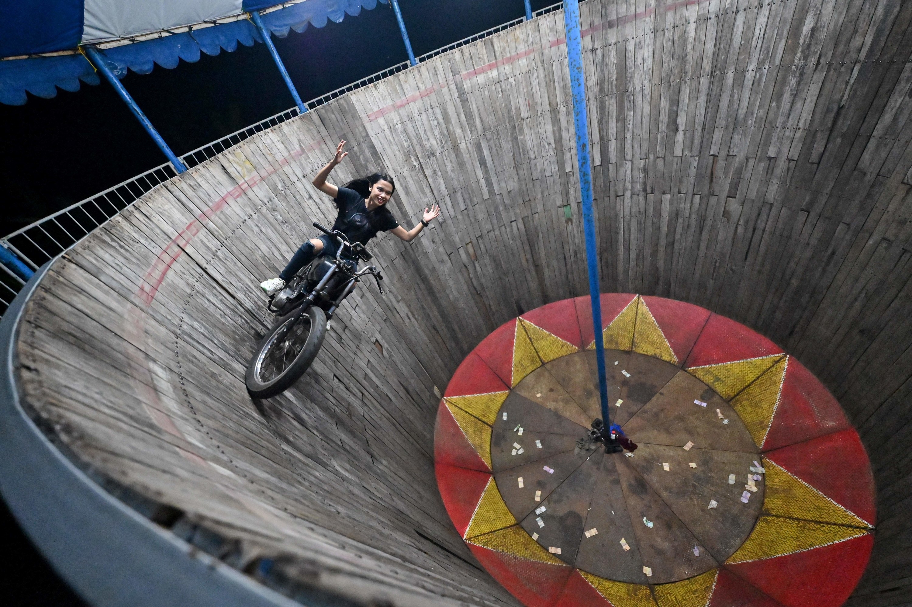 Karmila Purba, a young daredevil, rides the walls of the &quot;Well Of Death.&quot; She is smiling and throwing her hands in the air, vertical to the ground on a motorcycle.