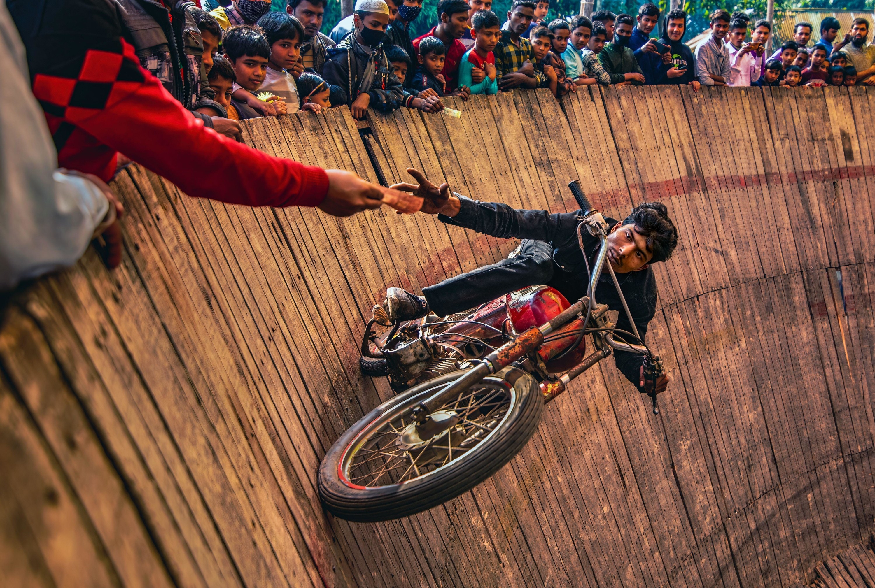 A man riding a motorcycle on the &quot;Well Of Death&quot; grabs a tip offered by an audience member as he rides past, vertically to the ground.