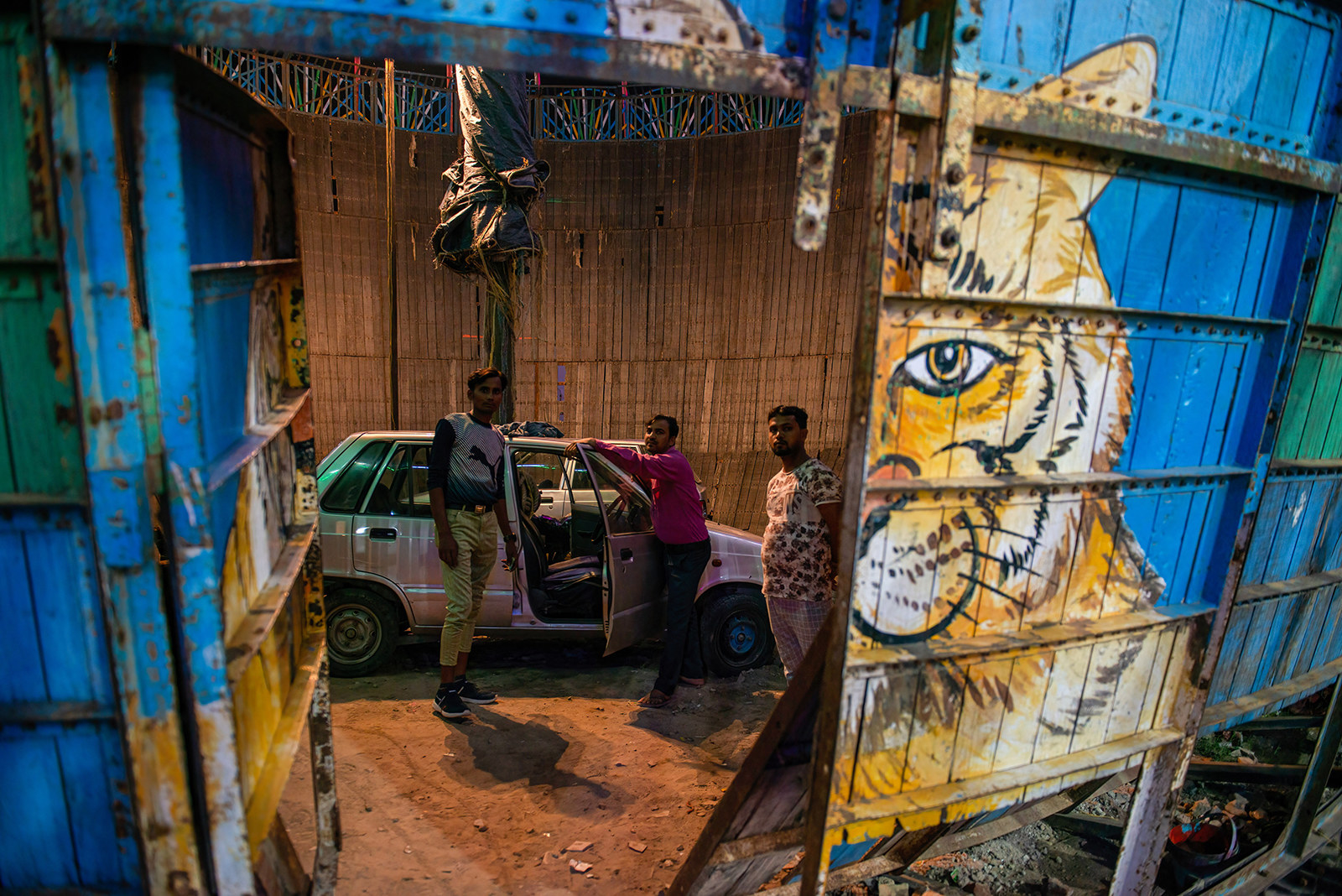 Three men stand in the &quot;Well of Death&quot; by a silver car with the doors open. The photography is taking the picture from outside of the structure through the door to the arena. There is a tiger painted on the outside wall.