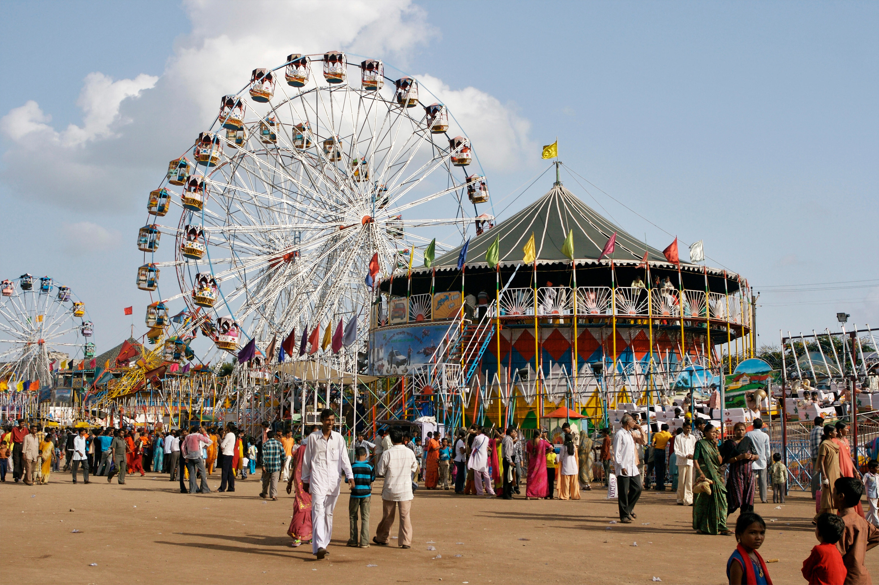 A fairgrounds that pictures the exterior of the Well Of Death. It is a tall cylinder where the audience stands at the top most section, looking down at the performers. The fairgrounds are bright and colorful.