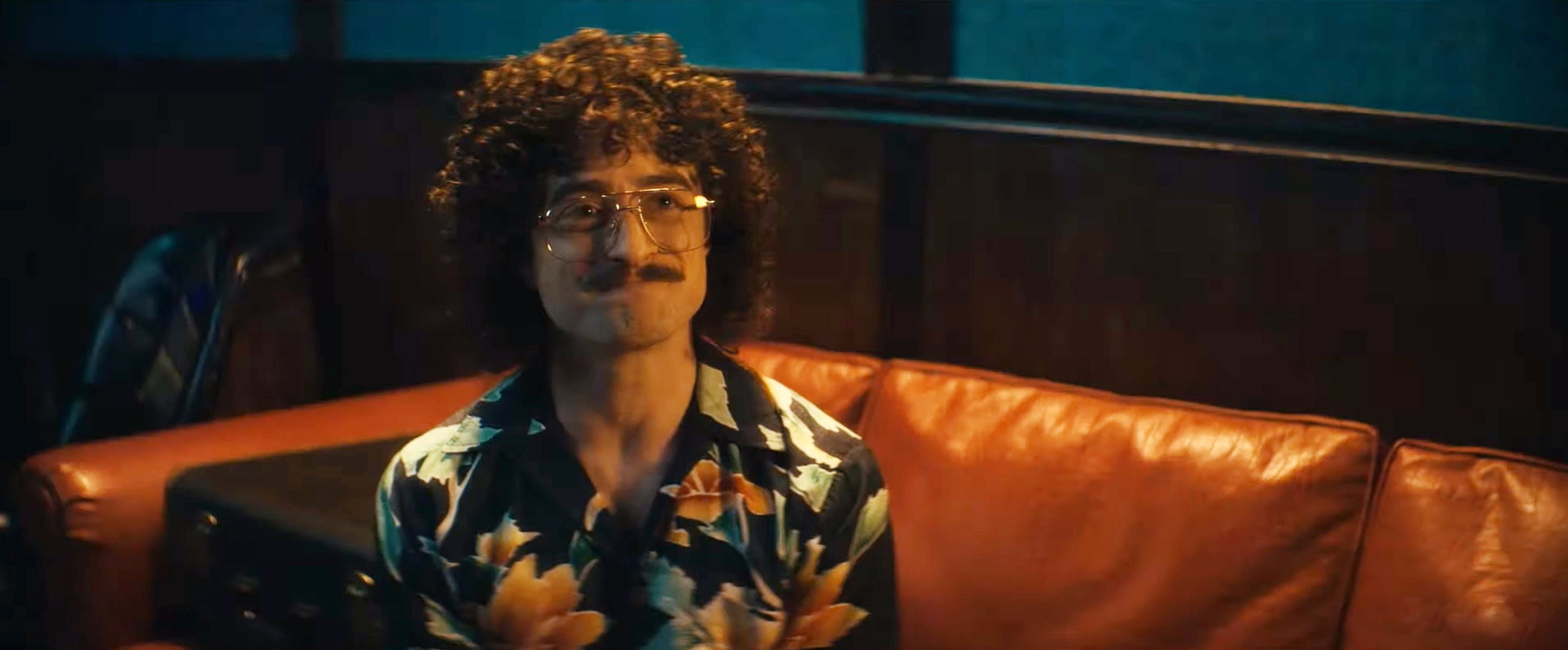 Daniel as Al sitting on a couch and rocking curly hair, a mustache, and large eyeglasses