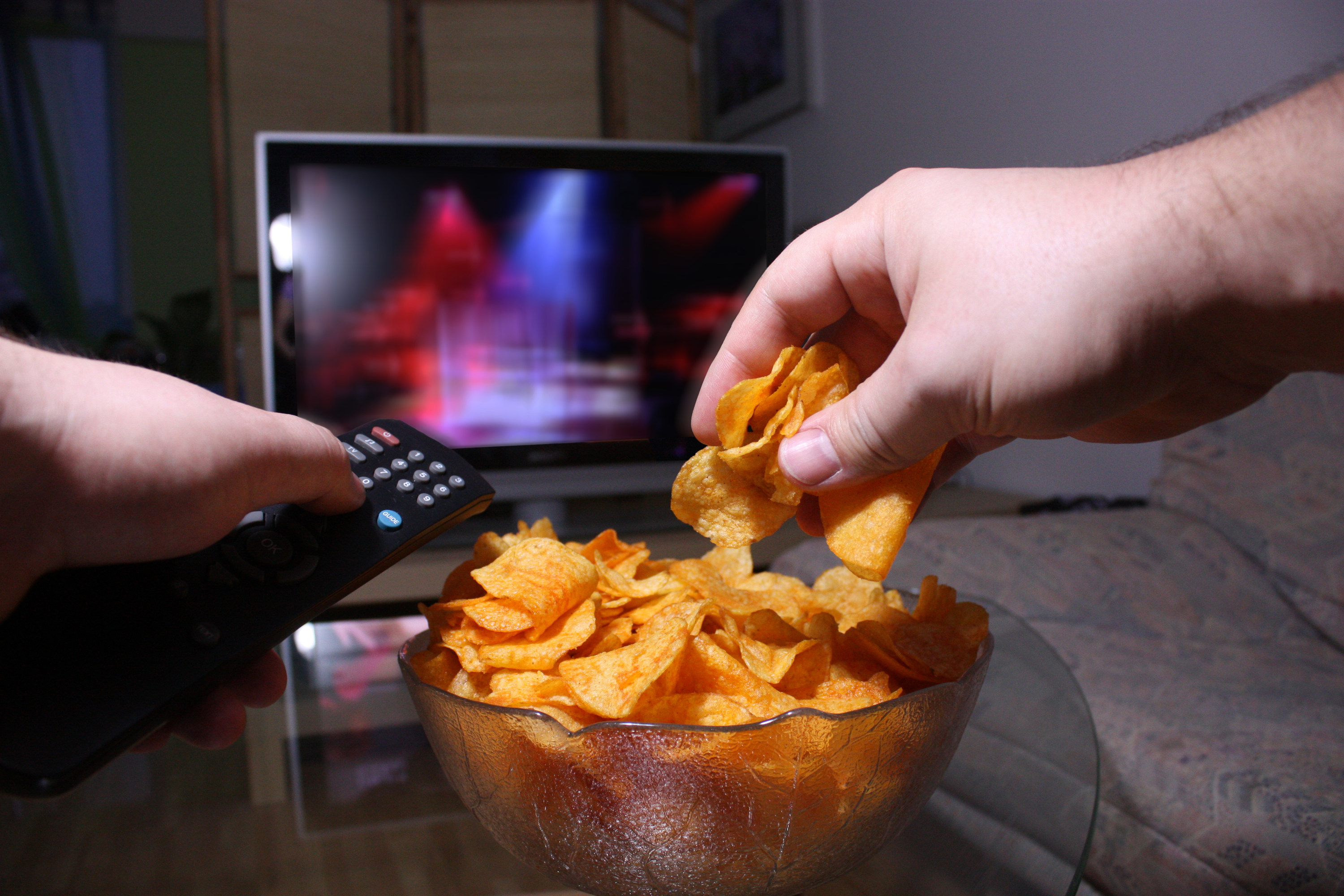 person eating a bowl of chips and turning on their tv with a remote control