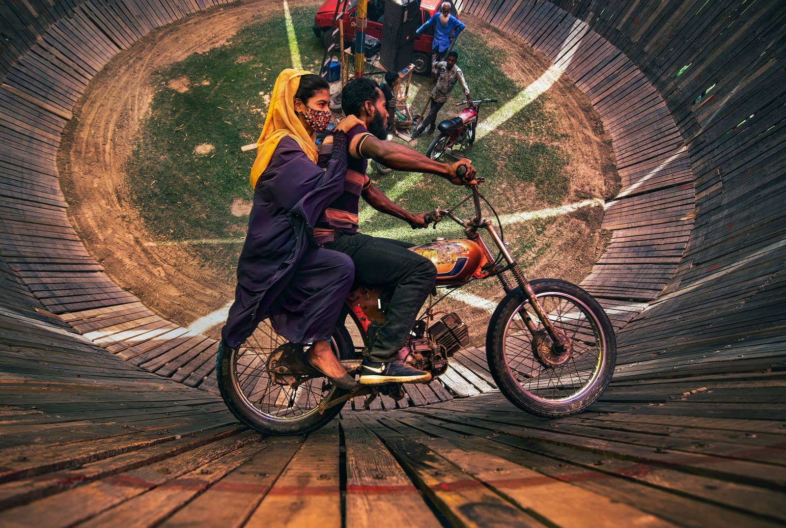 An aerial view looking into the &quot;Well Of Death,&quot; where a man and woman ride a motorcycle around a barrel shaped cylinder, nearly vertical to the ground.
