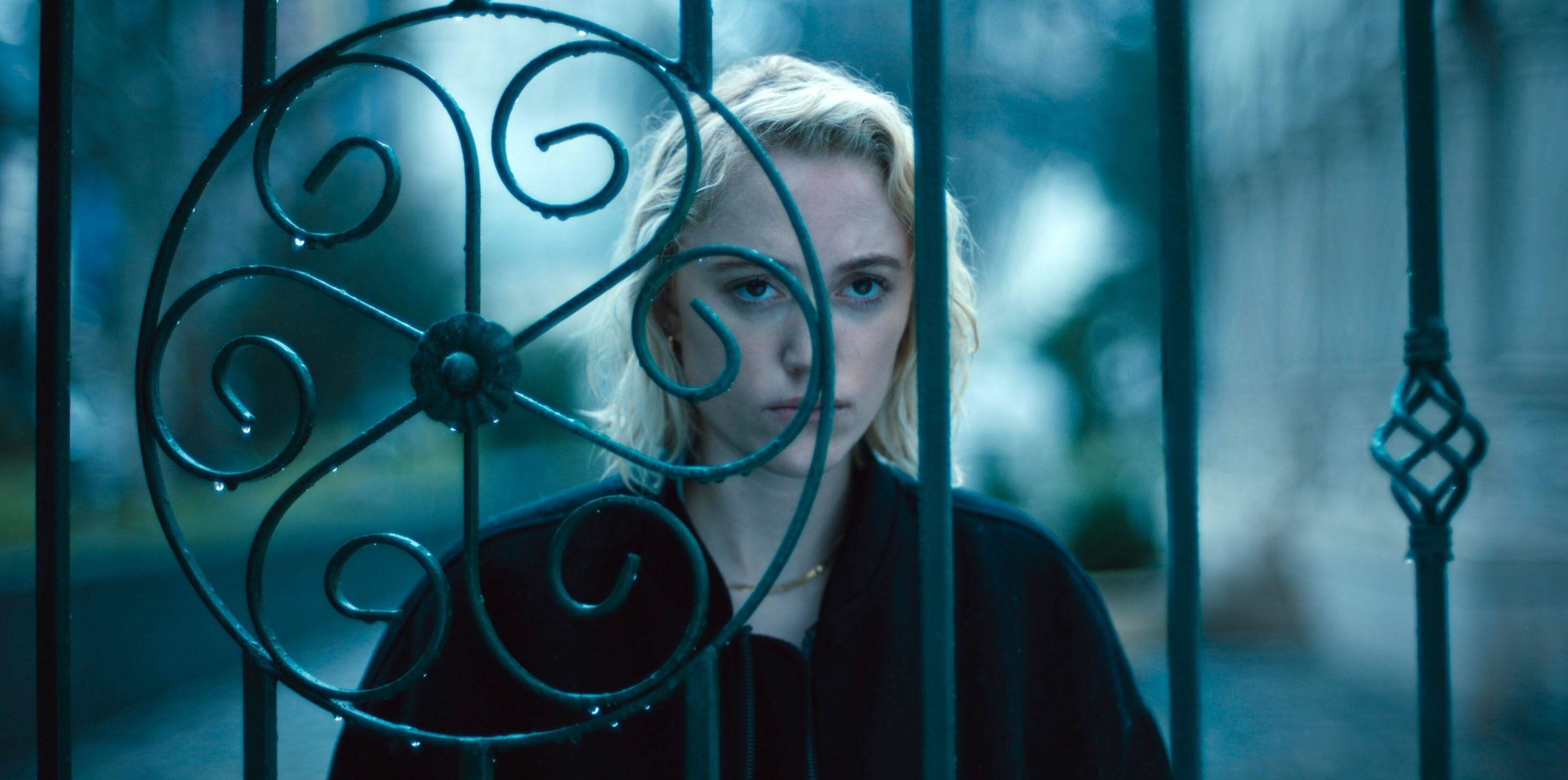 A young blonde woman stares at a stranger through an intricately designed fence in “Watcher”