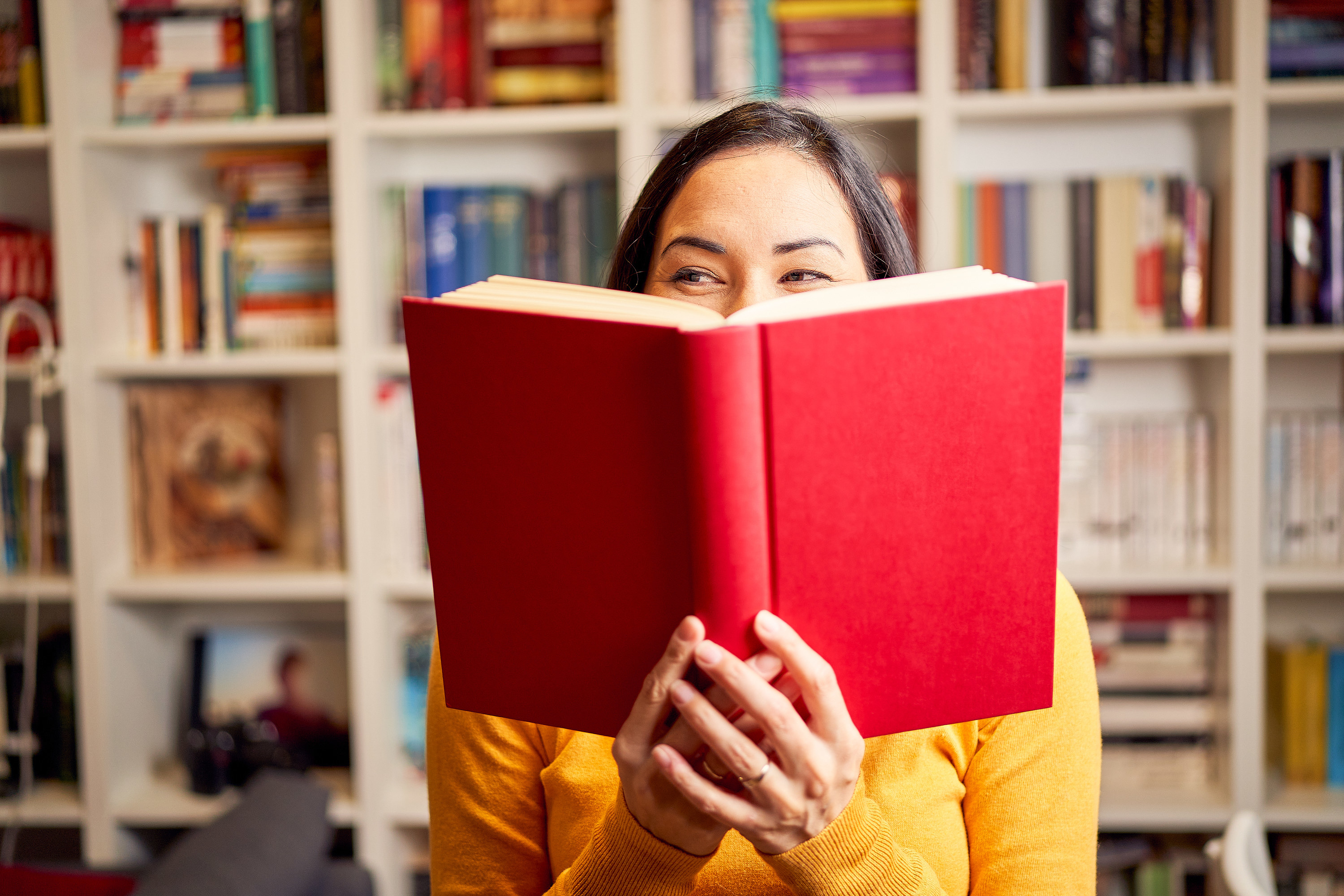 Smiling beautiful young female reading book standing against bookshelf at home