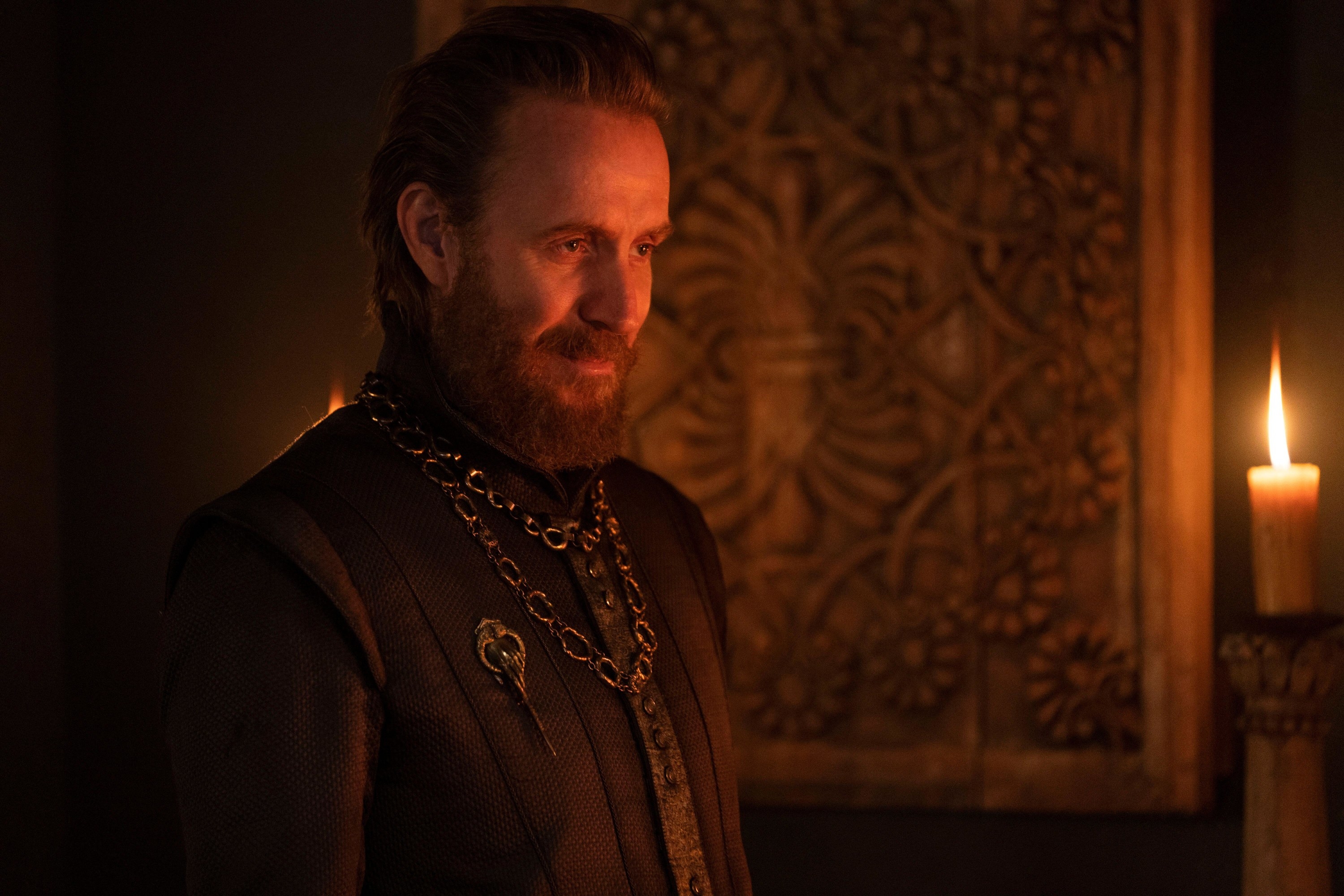 A bearded man in sleek clothing grins deceptively by candlelight