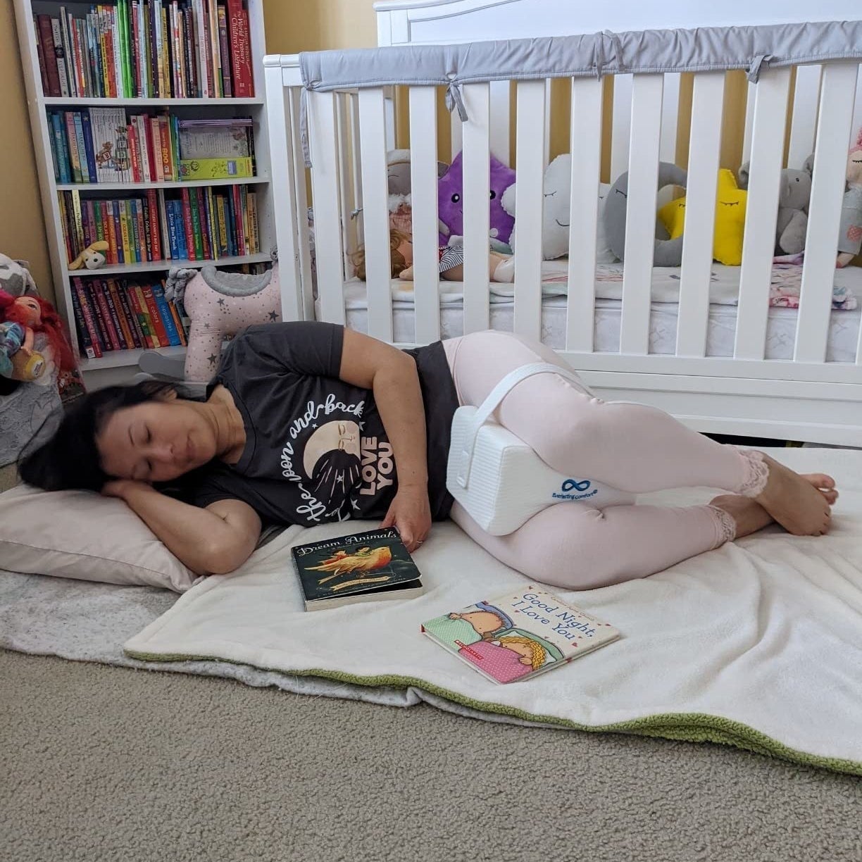 a reviewer sleeping on their side on the floor in front of a crib with the pillow between their knees