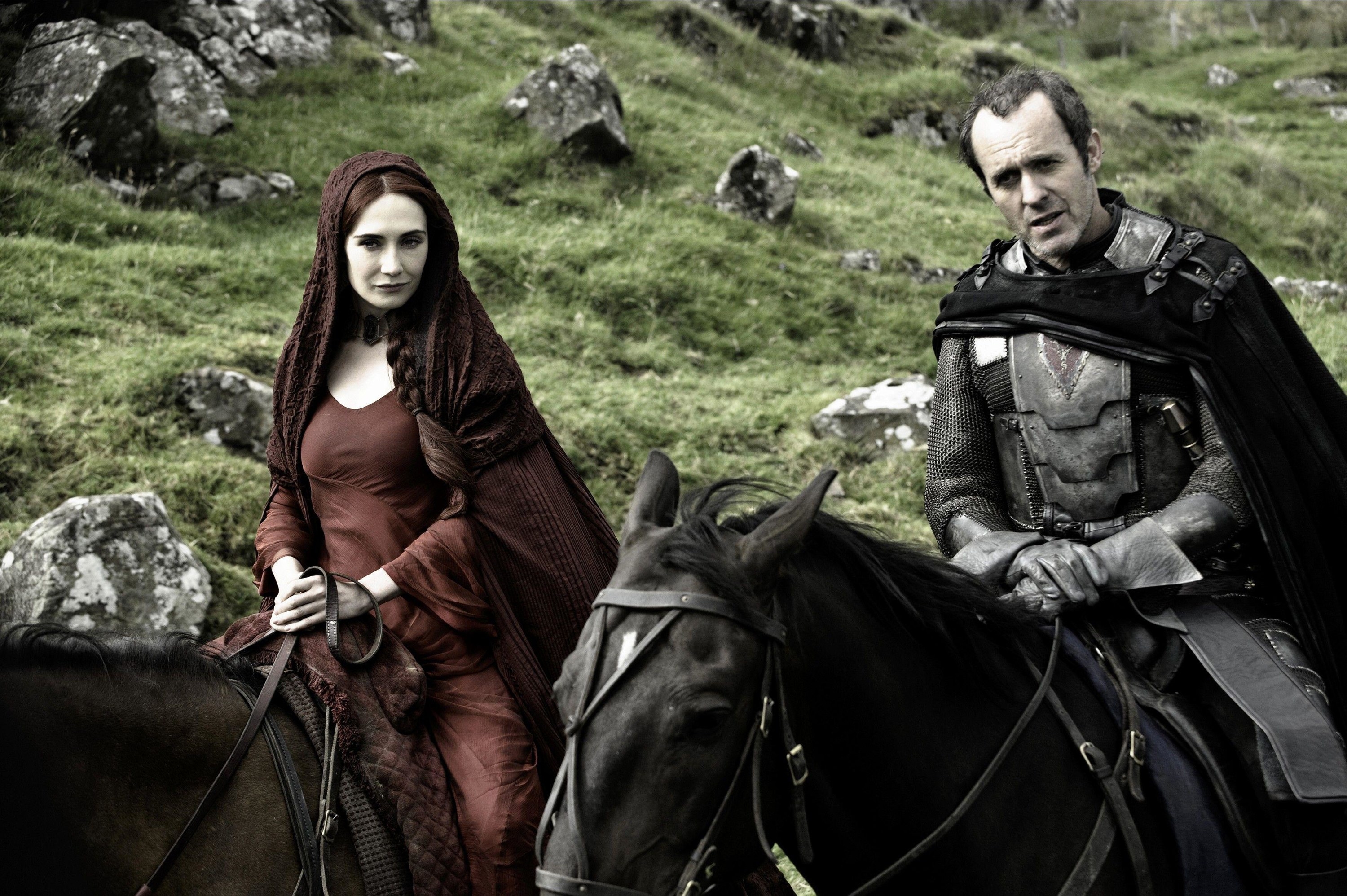 A woman in a red dress and cloak rides her horse alongside a man in all-black leathers and a cape.