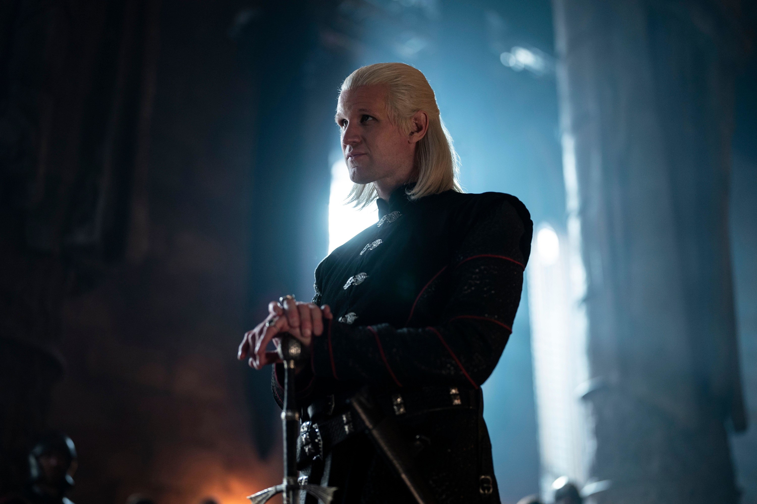 A blonde man stares disapprovingly in a great hall while placing his hands above his sword.