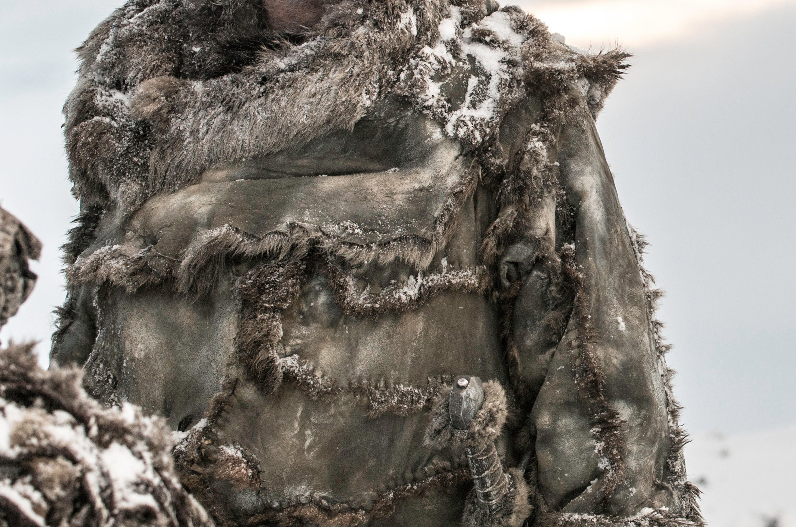 A man wearing a coat assembled from fur pelts observes a situation in a frozen tundra.