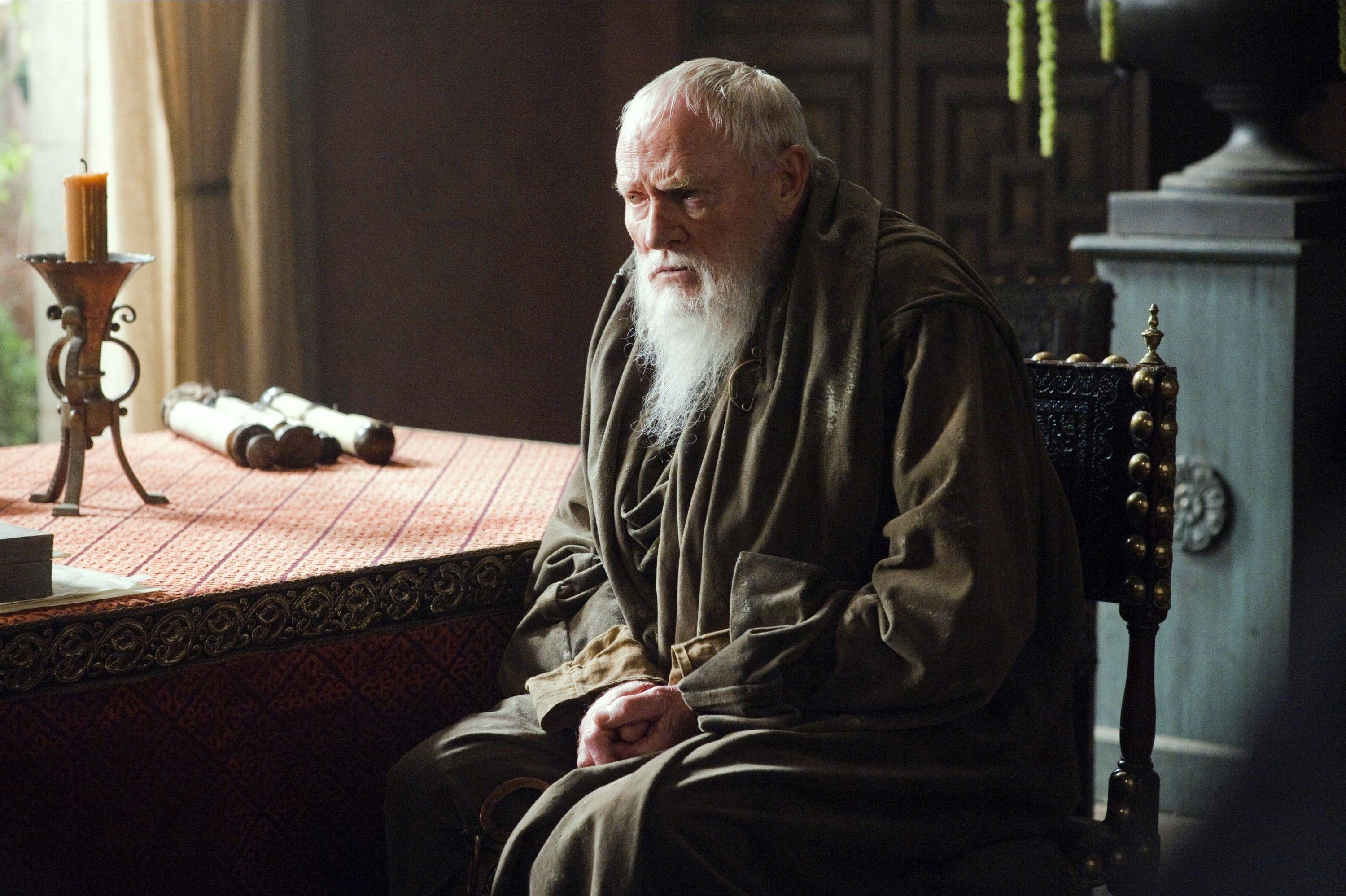 An old, bearded man in long robes sits at a table near some scrolls.