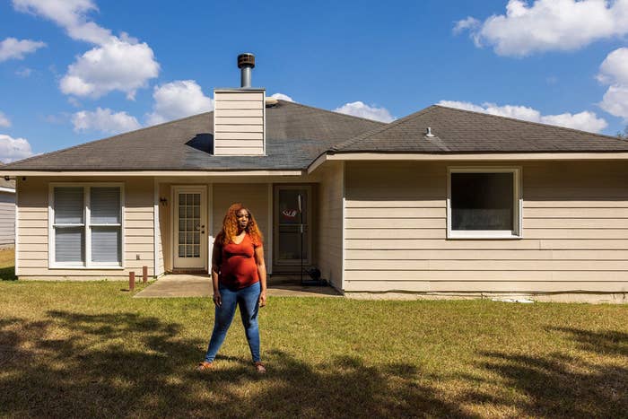 a woman with curly hair and a reddish orange t shirt stands in front of her house