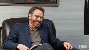 Bryan Cranston nodding his head and opening his arm while he sits in a chair