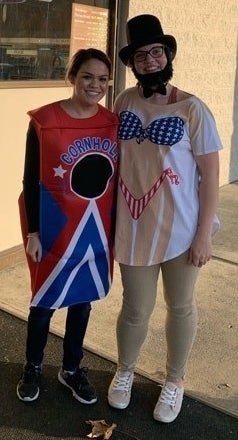 A person wearing a top hat, fake beard, bow tie, and a white T-shirt with a Stars and Stripes–themed bikini design, standing next to someone in a &quot;Cornhole&quot; costume