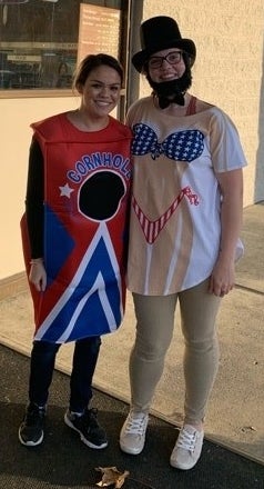 A person wearing a top hat, fake beard, bow tie, and a white T-shirt with a Stars and Stripes–themed bikini design, standing next to someone in a "Cornhole" costume