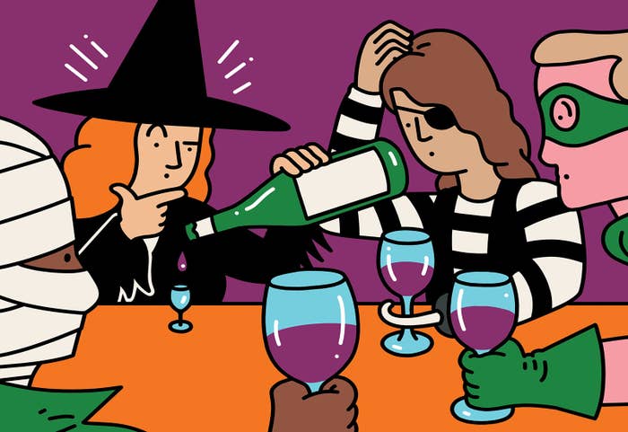 A group of friends sitting around a table, dressed in Halloween costumes. Three of the friends have full-sized wine glasses, but the fourth, dressed as a witch, is pouring one drop into a small glass