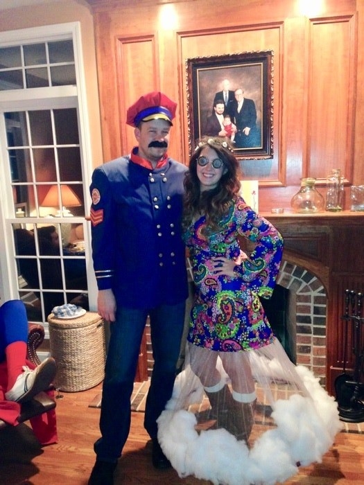 Person in jeans and a military jacket and hat and fake beard, and another in a psychedelic miniskirt with a long, cottony bottom and spiral glasses with flowers in her hair