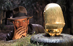 a gif of harrison ford as indiana jones looking at a golden idol and debating if he should take it