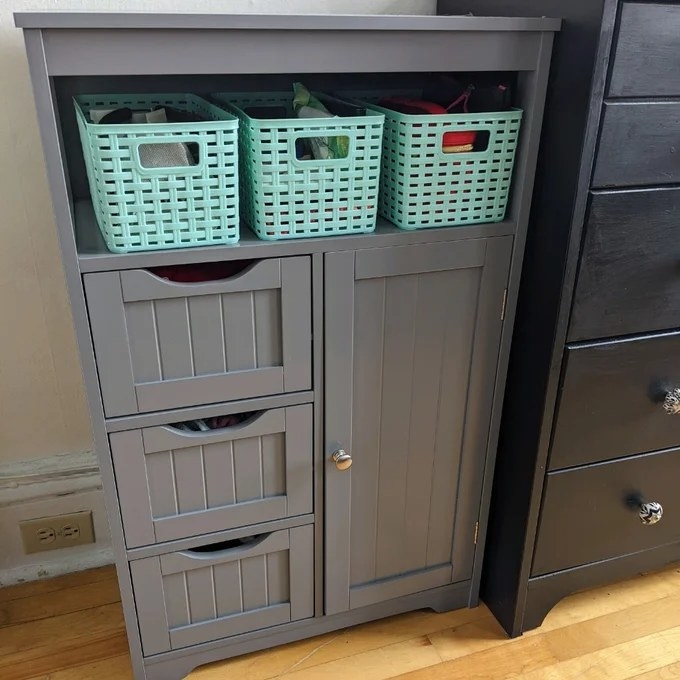 A gray freestanding cabinet with three drawers, one door and a rack holding three baskets