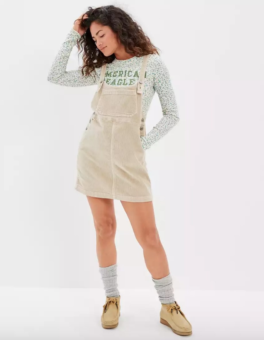 Model wearing beige corduroy overall mini dress over green sweater with gray socks and beige moccasin shoes