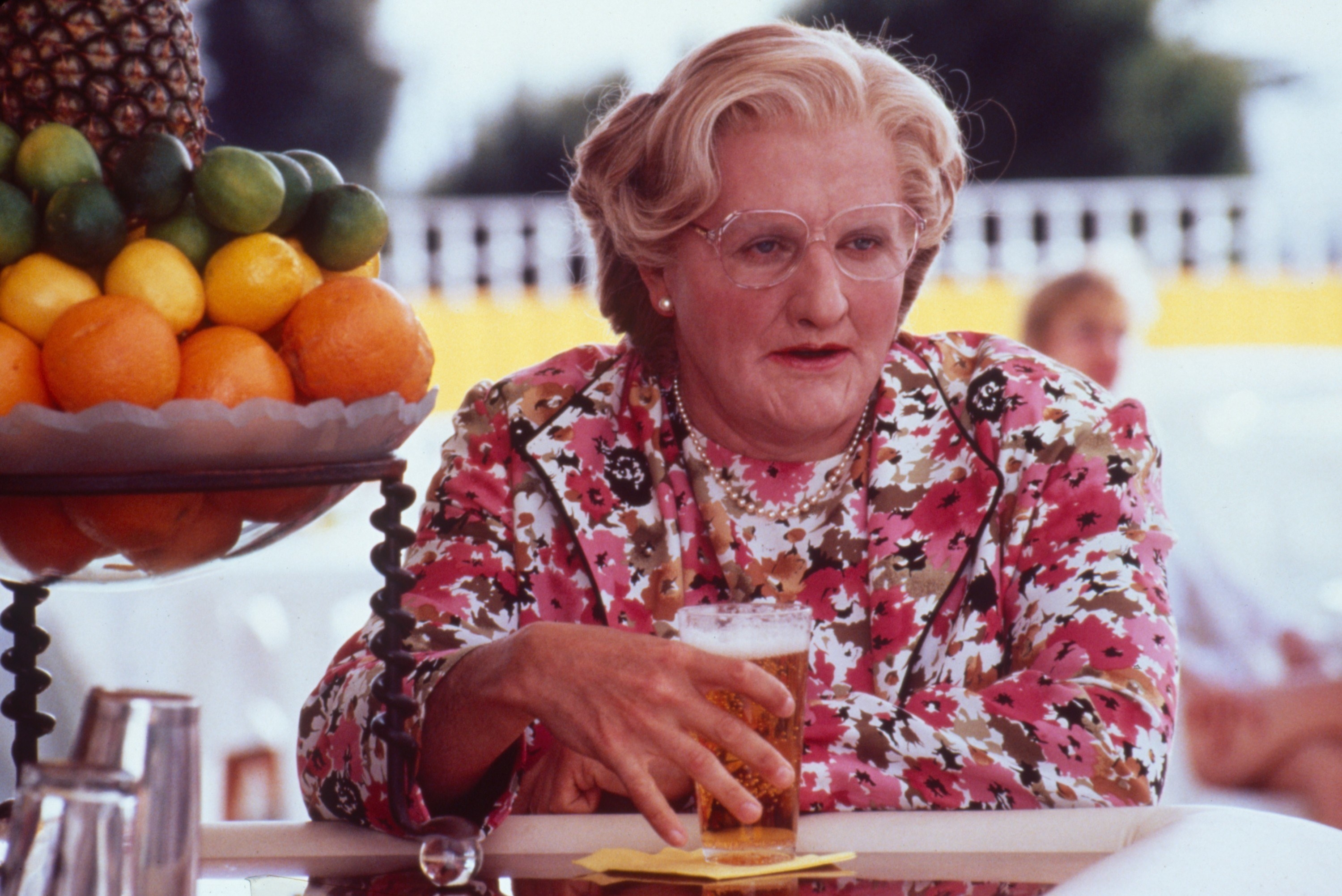 Mrs Doubtfire sitting next to a bowl of fruit holding a glass of beer