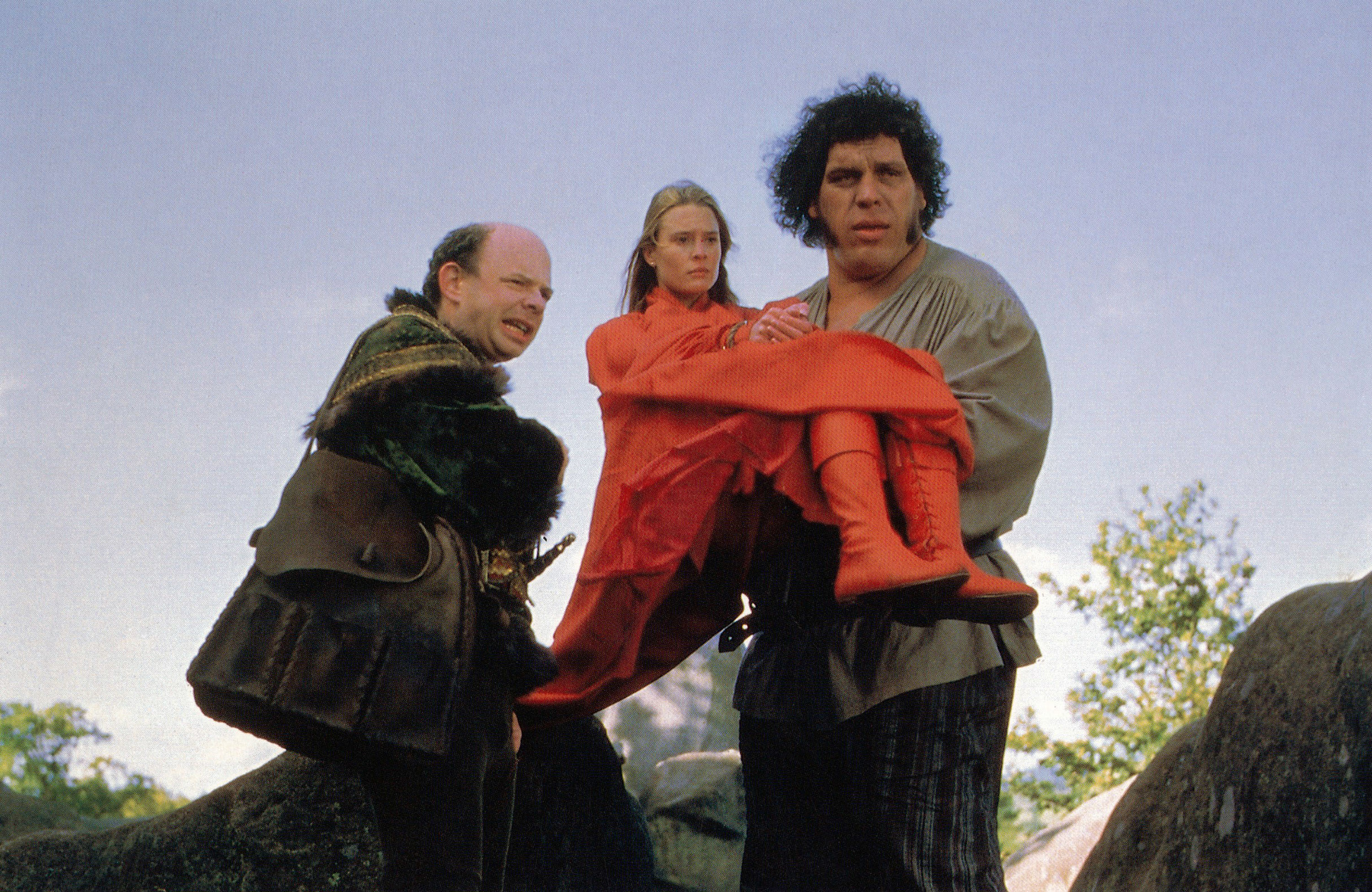 A large man holding a woman in his arms stands next to another man wearing a cape