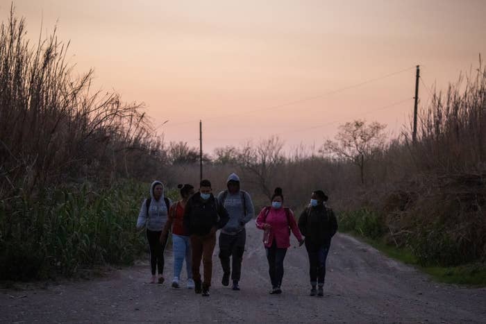 Six people, some wearing surgical masks, walk along a road with overgrown grass and trees on either side