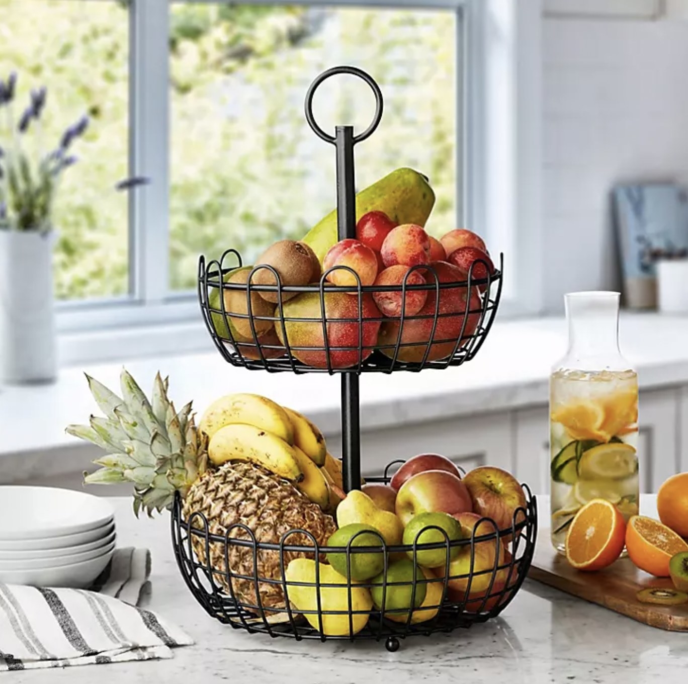 The fruit stand with pineapple, bananas, pears, and apples in larger bottom basket and plums, kiwis, and mangoes in top basket