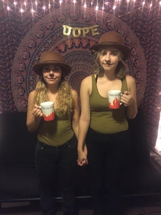 Two people holding hands, holding This Is Fine mugs, and wearing brown bowler-type hats