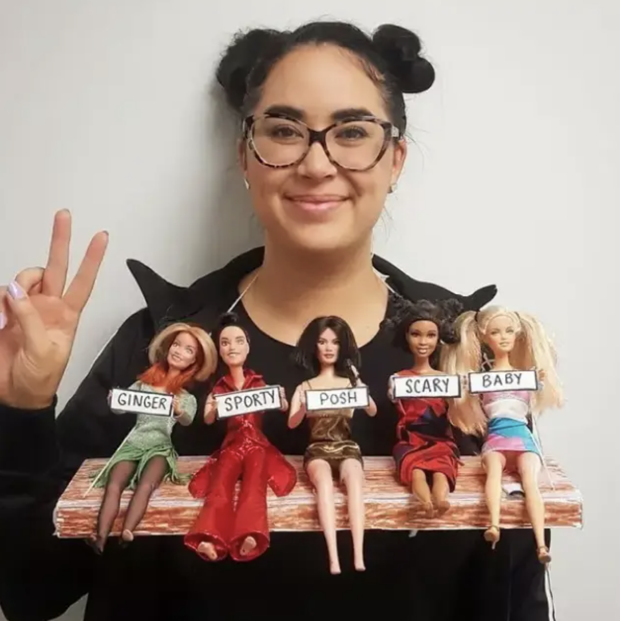 A woman wearing a piece of wood with five Barbie dolls with signs on them — Ginger, Sporty, Posh, Scary, and Baby — hanging from her neck and resting near her chest