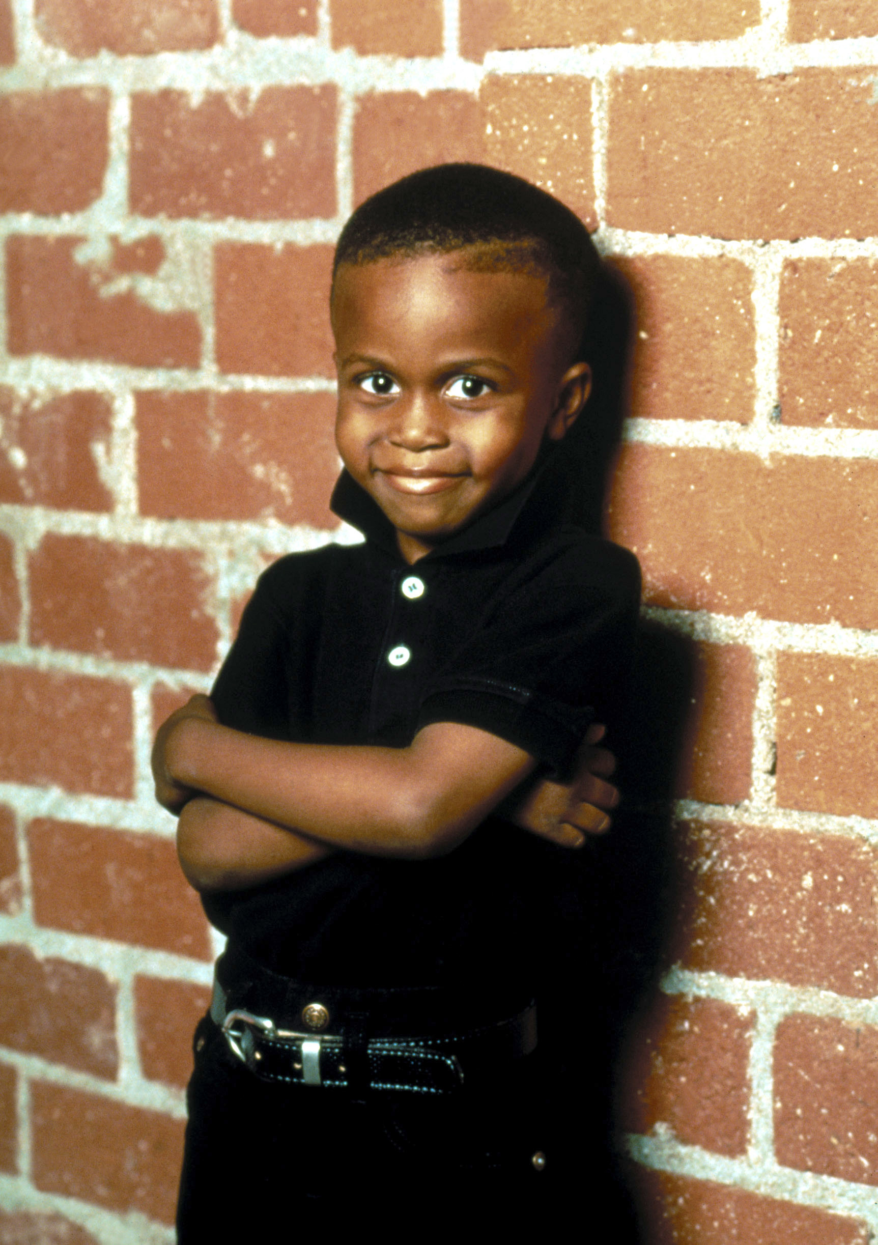 Ross as a little boy leaning against a brick wall with arms folded