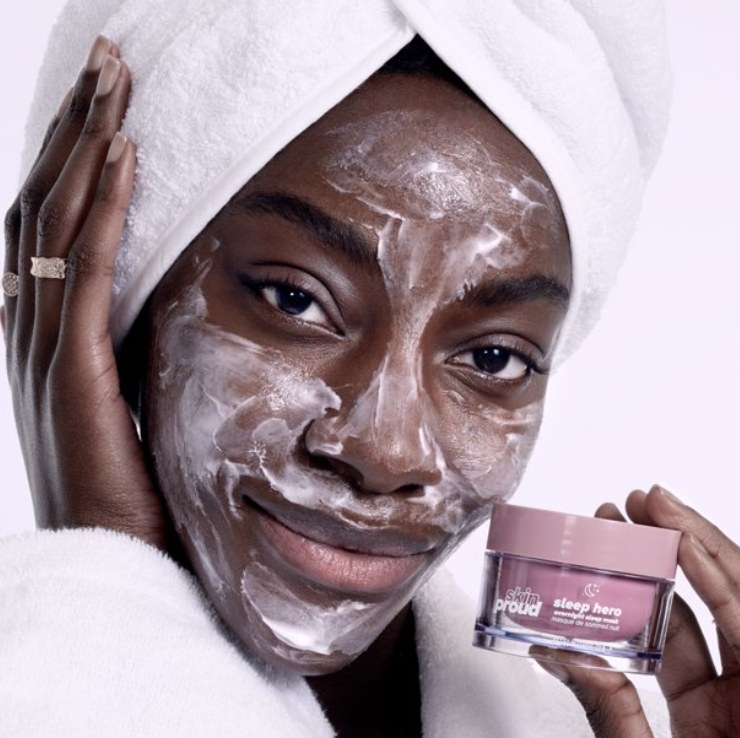 The model is wrapped in a robe and towel while holding the pink jar and the creamy mask is applied to their face