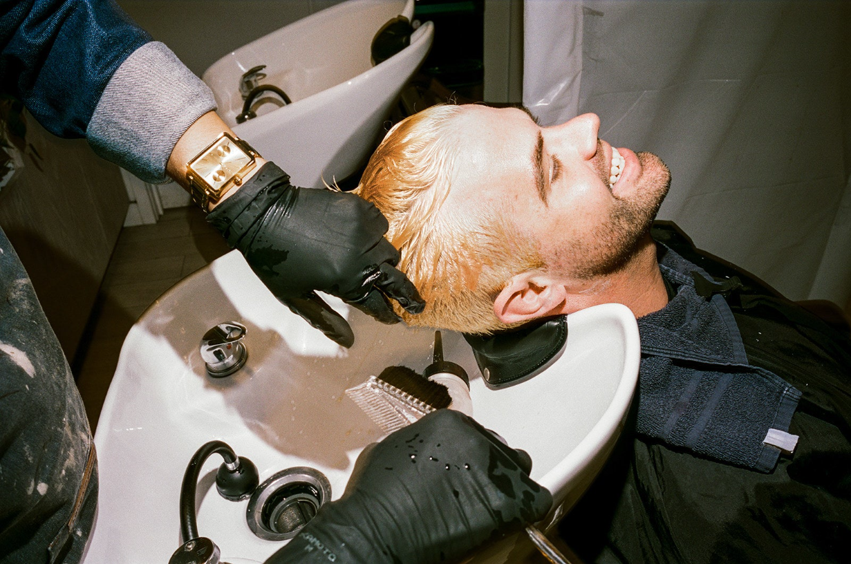 a man leans back into a sink as an unseen hair stylist wearing black gloves washes bleach out of his newly blonde hair